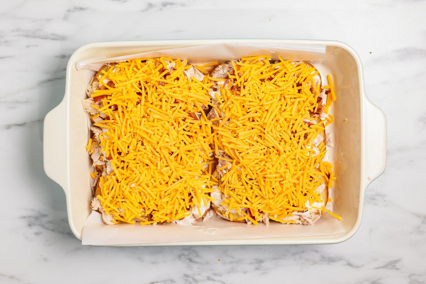 Shredded cheese added overtop of chicken, bacon, and ranch sliders in a baking dish