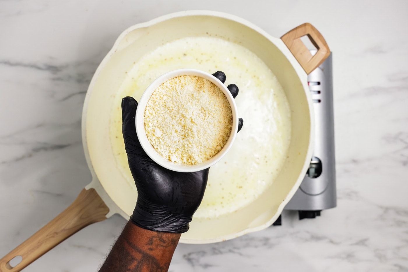 Hand holding a bowl of parmesan cheese over a skillet of garlic cream sauce