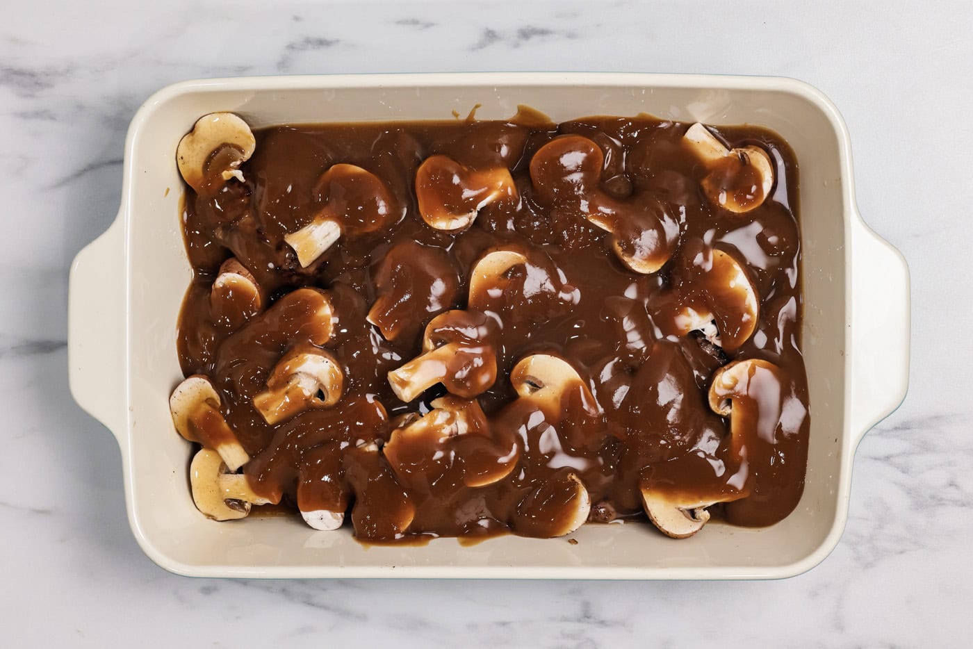 brown gravy poured over mushrooms and patties in a baking dish
