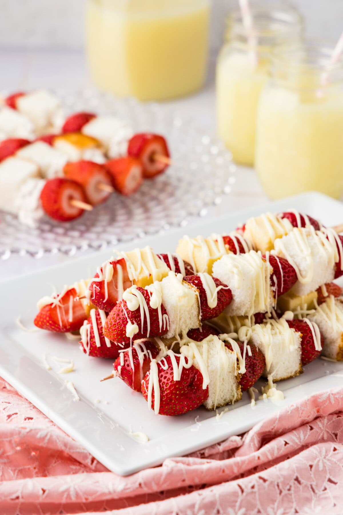 Strawberry Shortcake Kabobs on a platter drizzled with white chocolate, and more kabobs in back with whipped cream