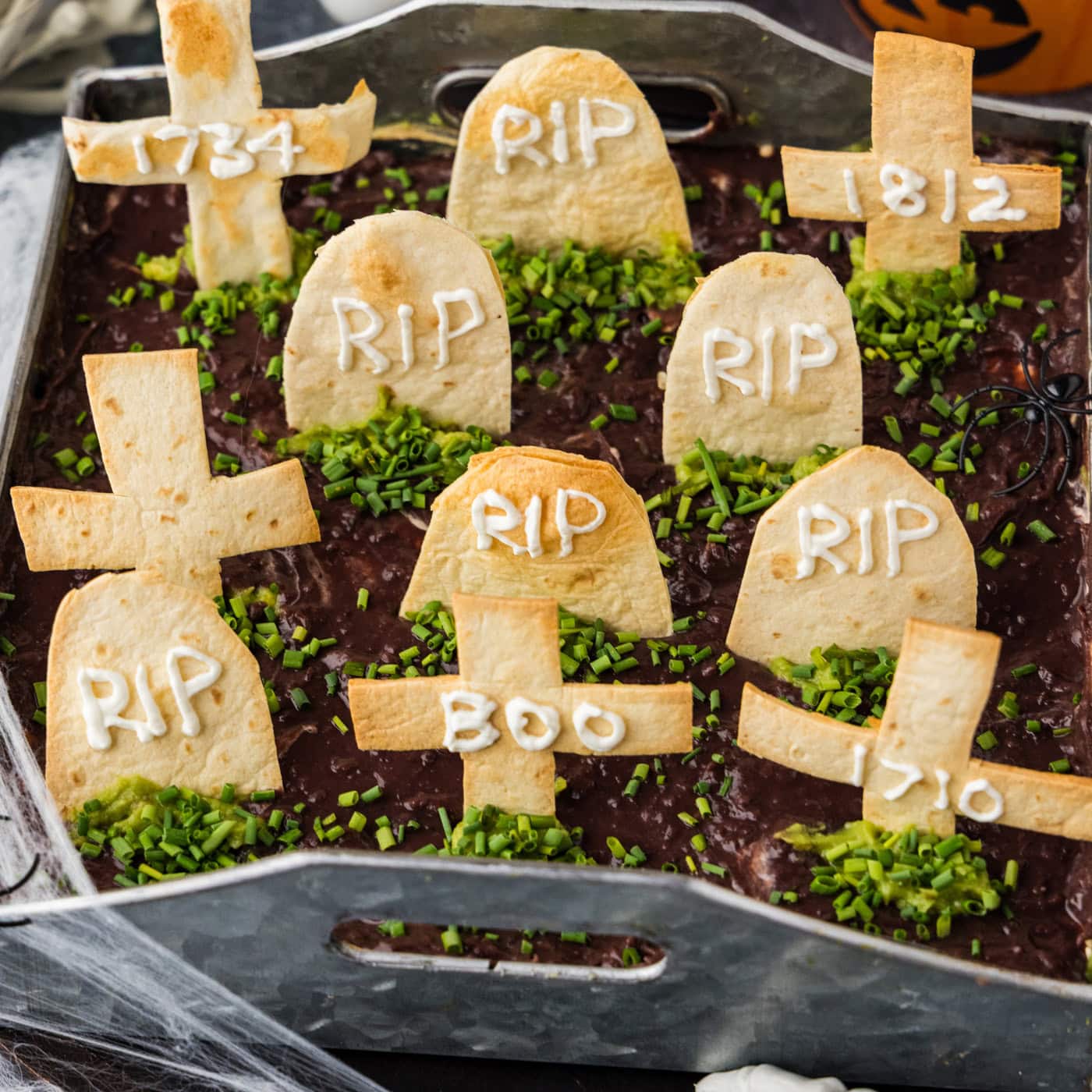 Graveyard taco dip in a square pan with tortilla tombstones standing in black refried bean dirt and chive grass