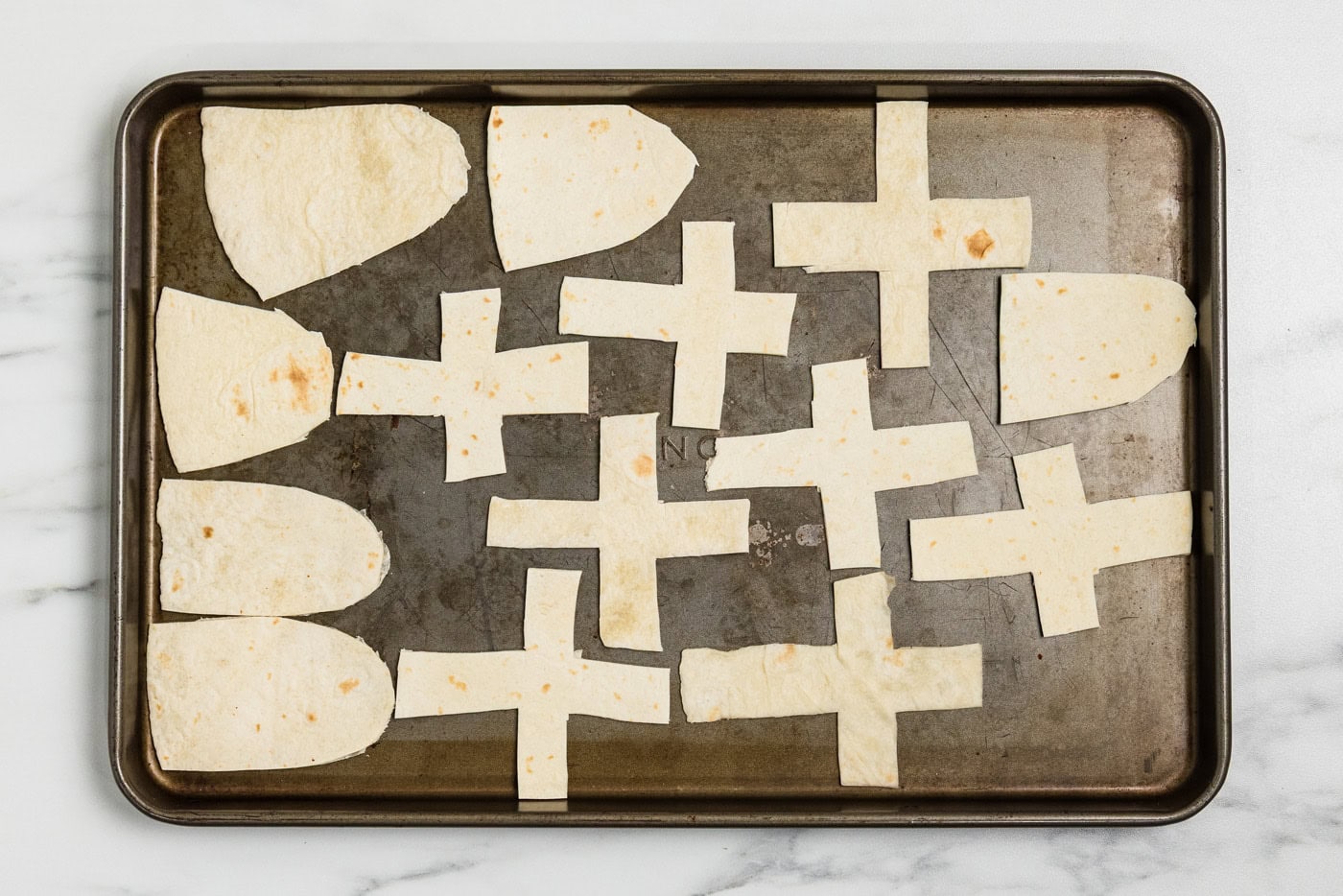 Flour tortillas cut into tombstone and cross shapes arranged on a baking sheet before toasting