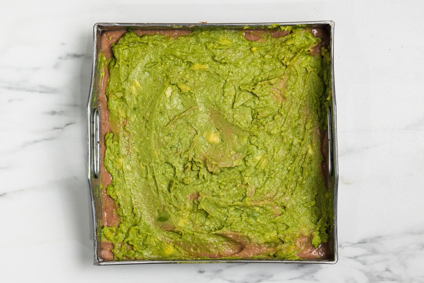 Guacamole spread over the refried bean layer in a square baking pan