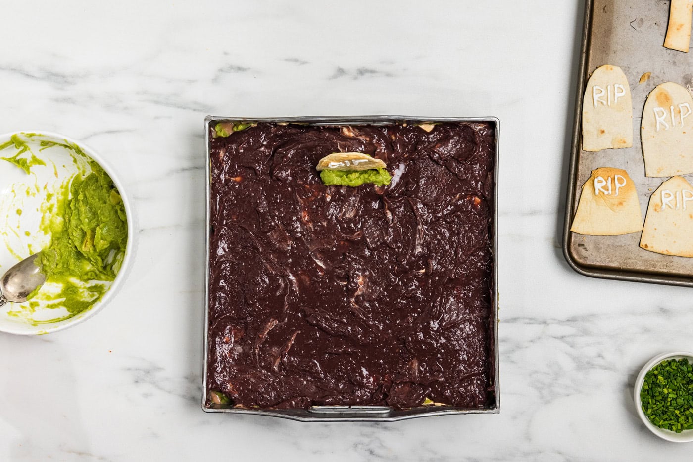 A tortilla tombstone being pushed into the black bean layer with reserved guacamole spooned at its base