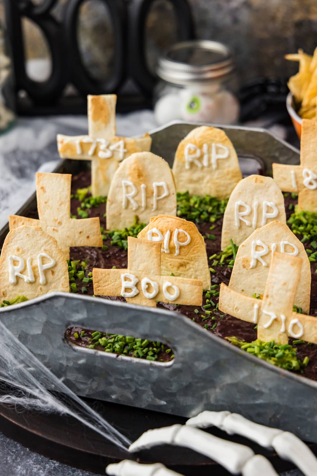 Graveyard taco dip topped with tortilla tombstones and chive grass, styled with cobwebs and a skeleton hand