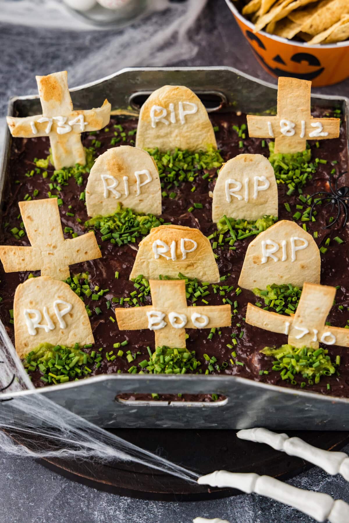 Graveyard taco dip with tortilla tombstones and chives next to a bowl of tortilla chips in a jack-o-lantern bowl