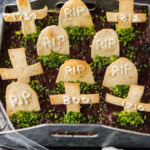Graveyard taco dip with tortilla tombstones and chives next to a bowl of tortilla chips in a jack-o-lantern bowl
