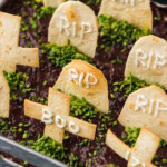 Close up of tortilla tombstones reading RIP and BOO standing in black refried beans with chopped chives