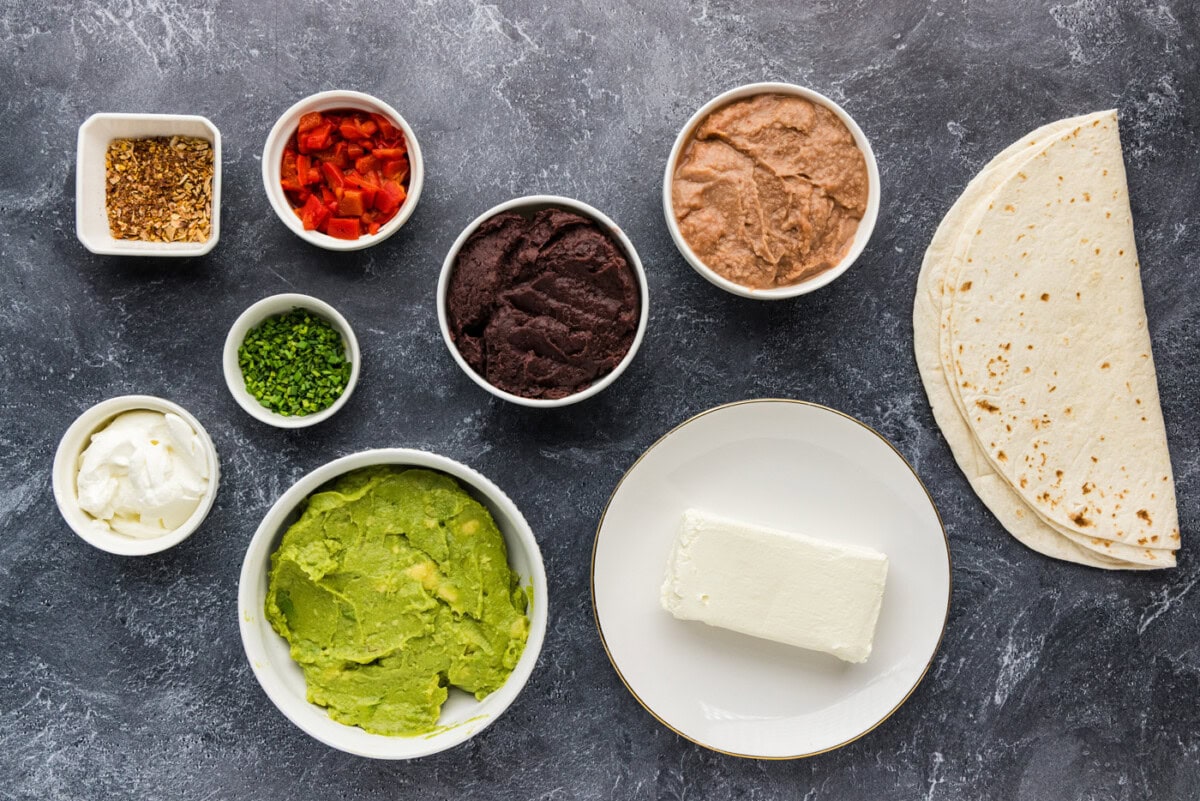 Ingredients for graveyard taco dip including refried beans, black refried beans, guacamole, cream cheese, sour cream, taco seasoning, roasted red peppers, chives, and flour tortillas