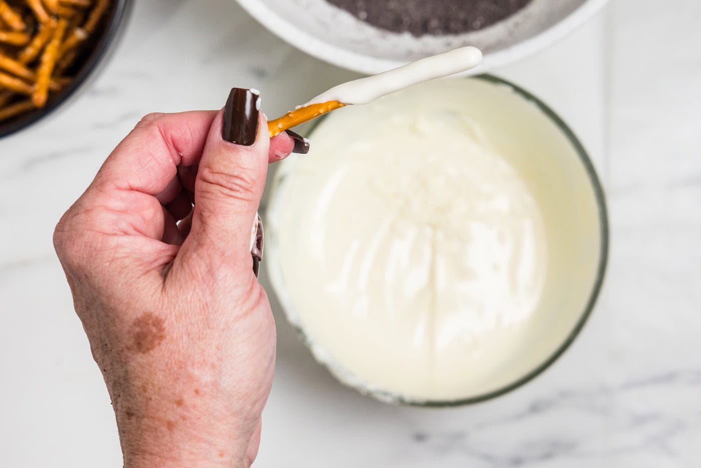 Melted white chocolate in a bowl with a hand ver it holding a dipped pretzel stick