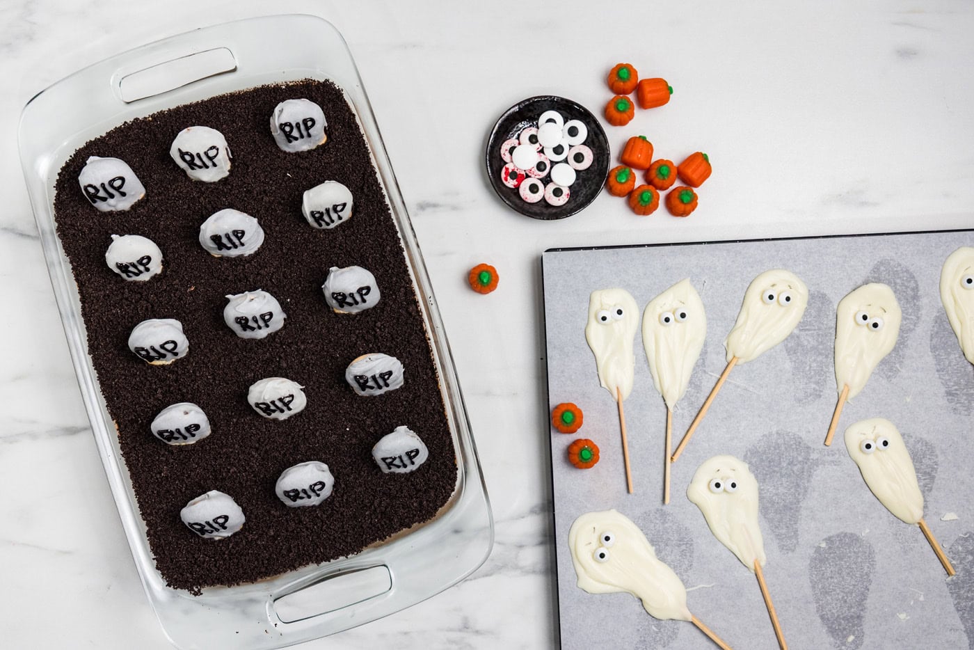 Cookie tombstones pressed into the Oreo dirt with candy eyeballs, candy pumpkins and finished ghost pops waiting alongside