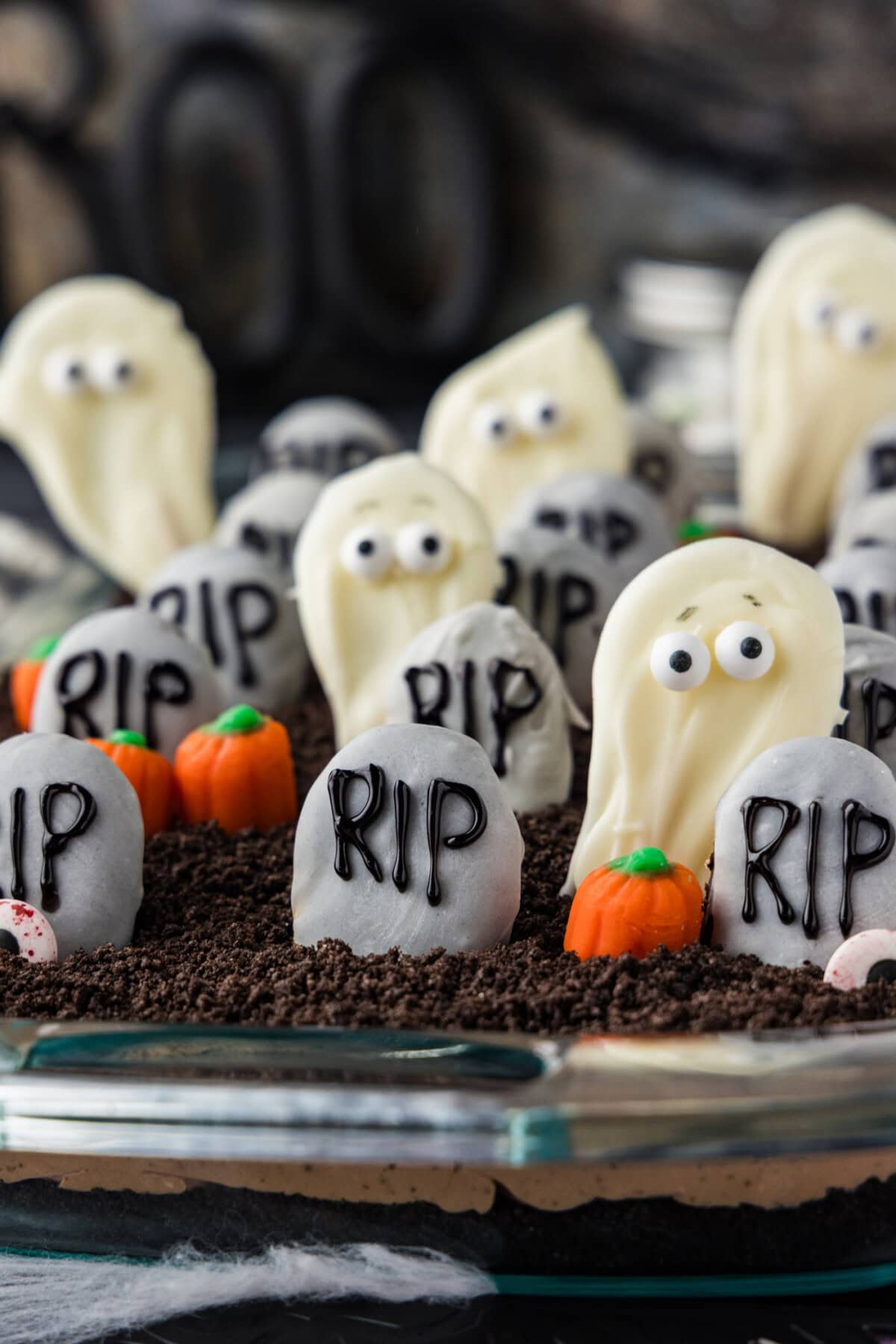 Cookie tombstones and white chocolate ghosts standing in Oreo crumb dirt with candy pumpkins between them