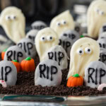 Cookie tombstones and white chocolate ghosts standing in Oreo crumb dirt with candy pumpkins between them
