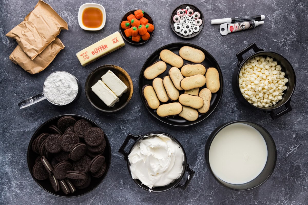 Oreos, cream cheese, butter, powdered sugar, milk, Cool Whip, Milano cookies, white chocolate chips, candy pumpkins and candy eyeballs arranged on a gray surface