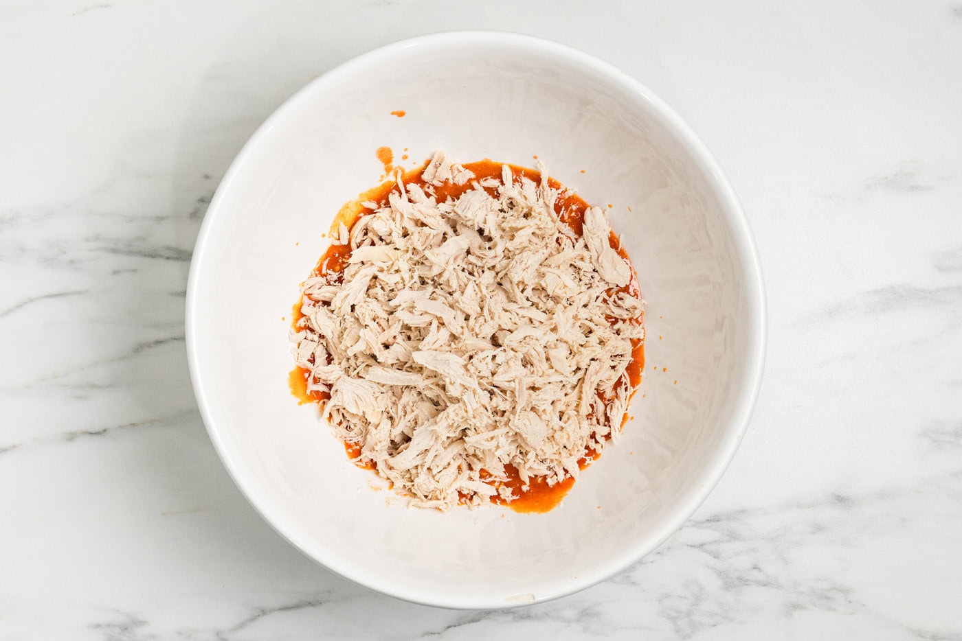 Shredded chicken added to the buffalo sauce in a white mixing bowl