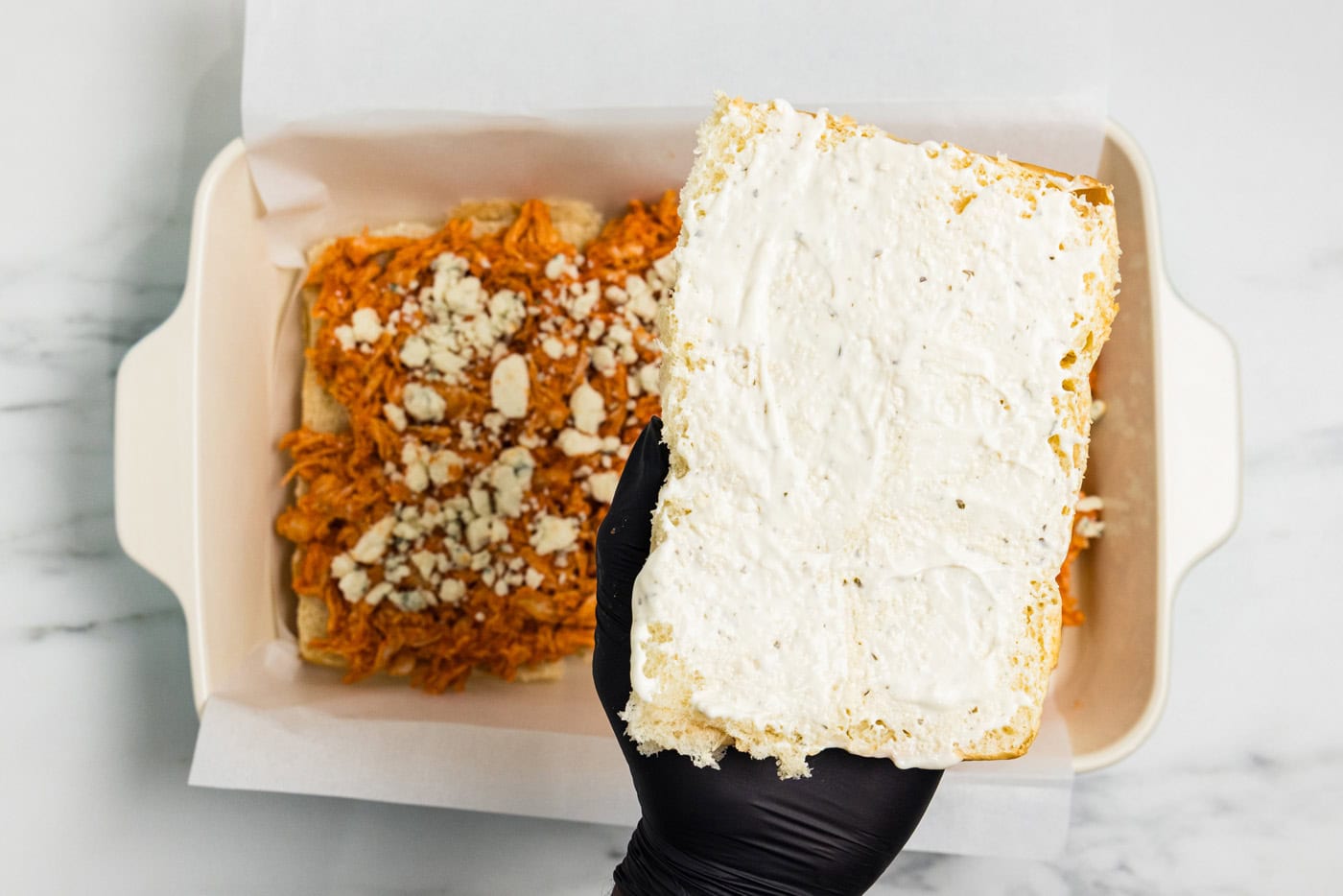 Hand holding the top sheet of Hawaiian rolls spread with blue cheese dressing over the buffalo chicken