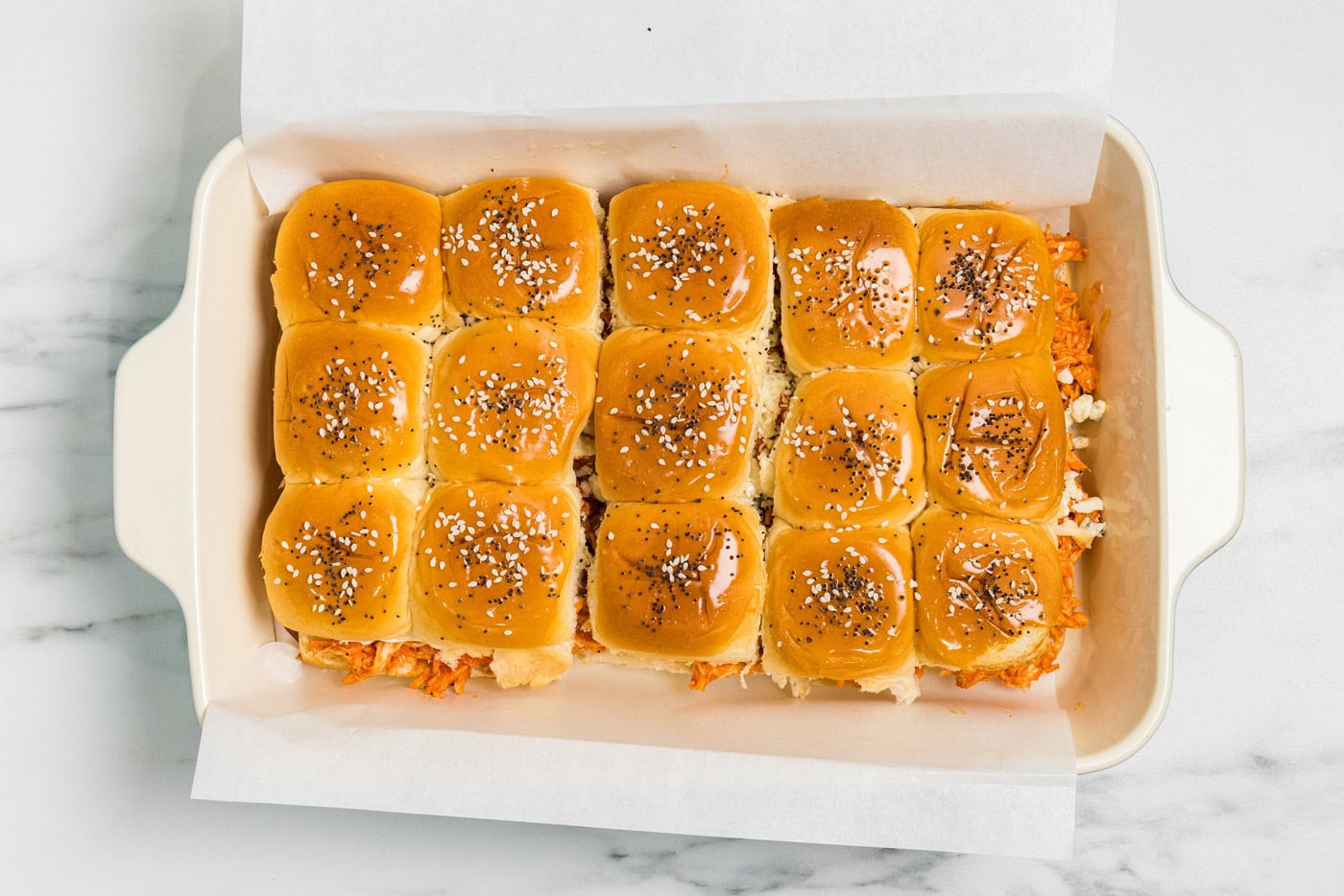 Assembled buffalo chicken sliders brushed with butter and topped with sesame and poppy seeds before baking