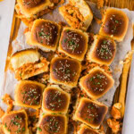 Overhead of buffalo chicken sliders with seeded tops on a wooden board with hot sauce and blue cheese dressing on the side