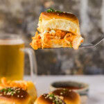 Spatula lifting a buffalo chicken slider with a glass of beer in the background