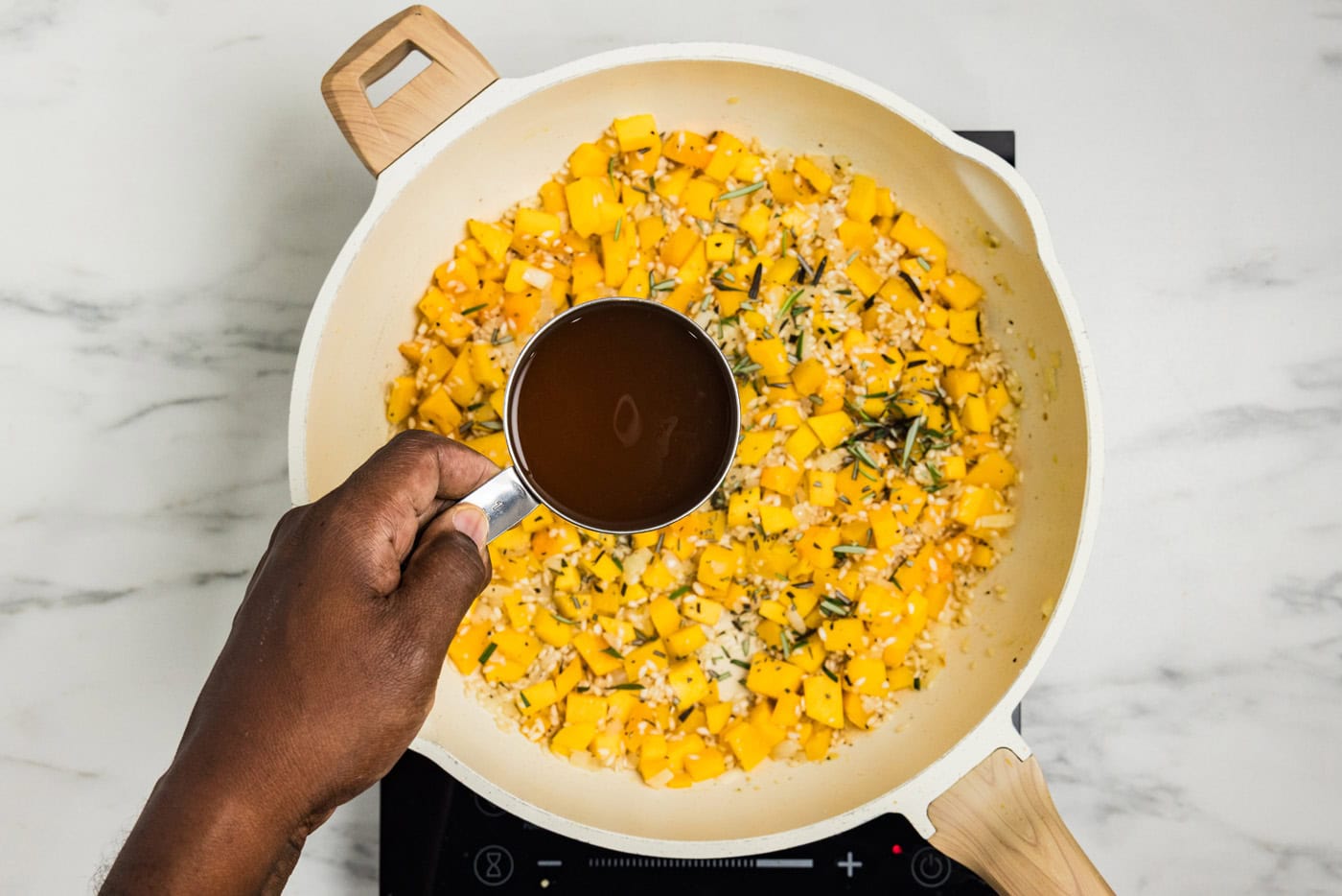 Warm vegetable broth being poured from a measuring cup into the butternut squash and rice in the skillet.