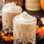 Two pumpkin spice mudslides topped with whipped cream and a dusting of spice beside a pumpkin and fall leaves