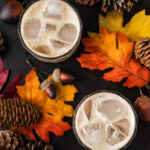 Overhead of two pumpkin spice mudslides over ice on a dark board with fall leaves, pinecones and acorns