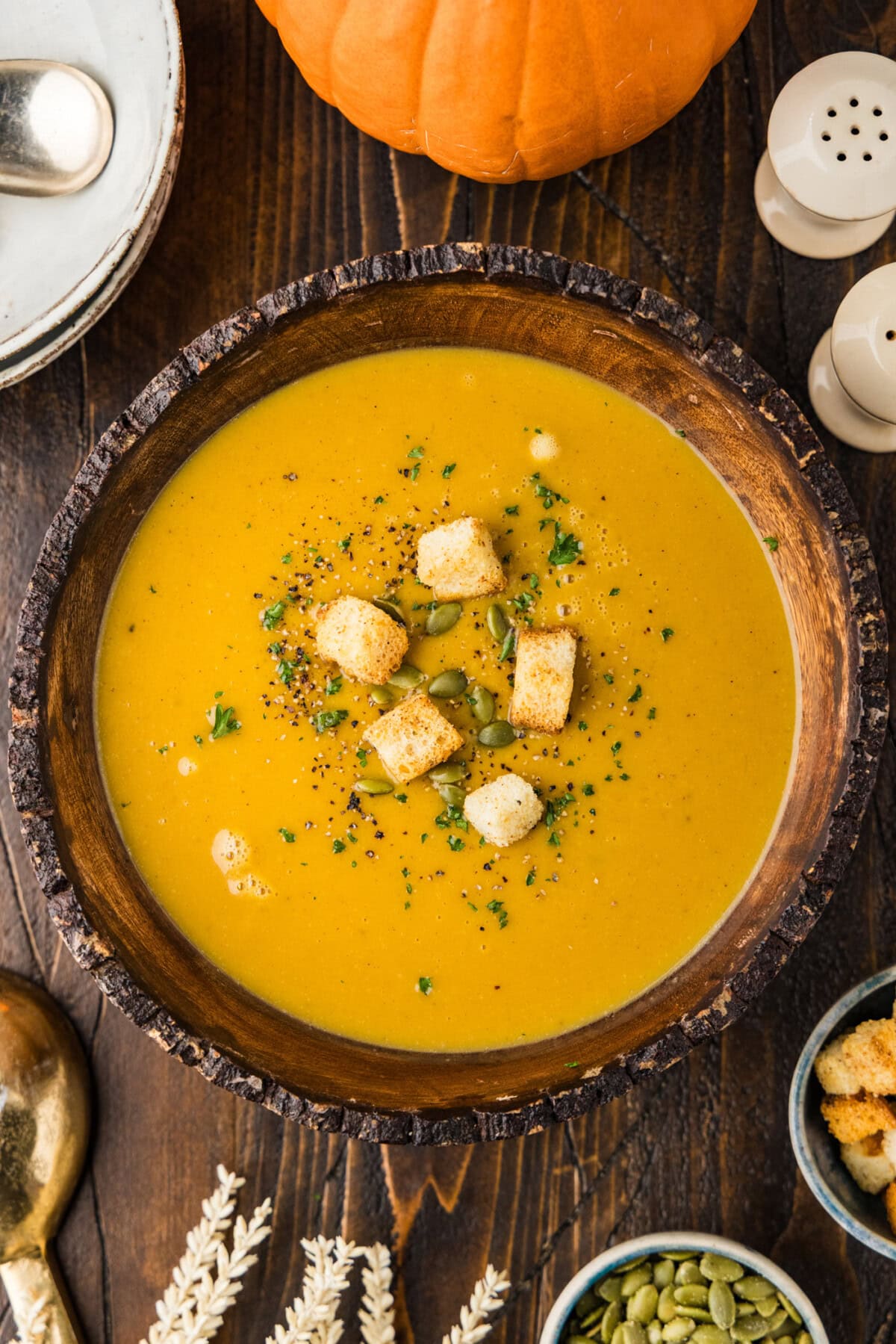 Overhead view of creamy roasted pumpkin soup in a wooden bowl topped with croutons, pepitas, parsley and black pepper, with a sugar pumpkin nearby.