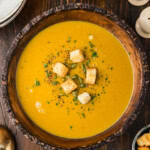 Overhead view of creamy roasted pumpkin soup in a wooden bowl topped with croutons, pepitas, parsley and black pepper, with a sugar pumpkin nearby.