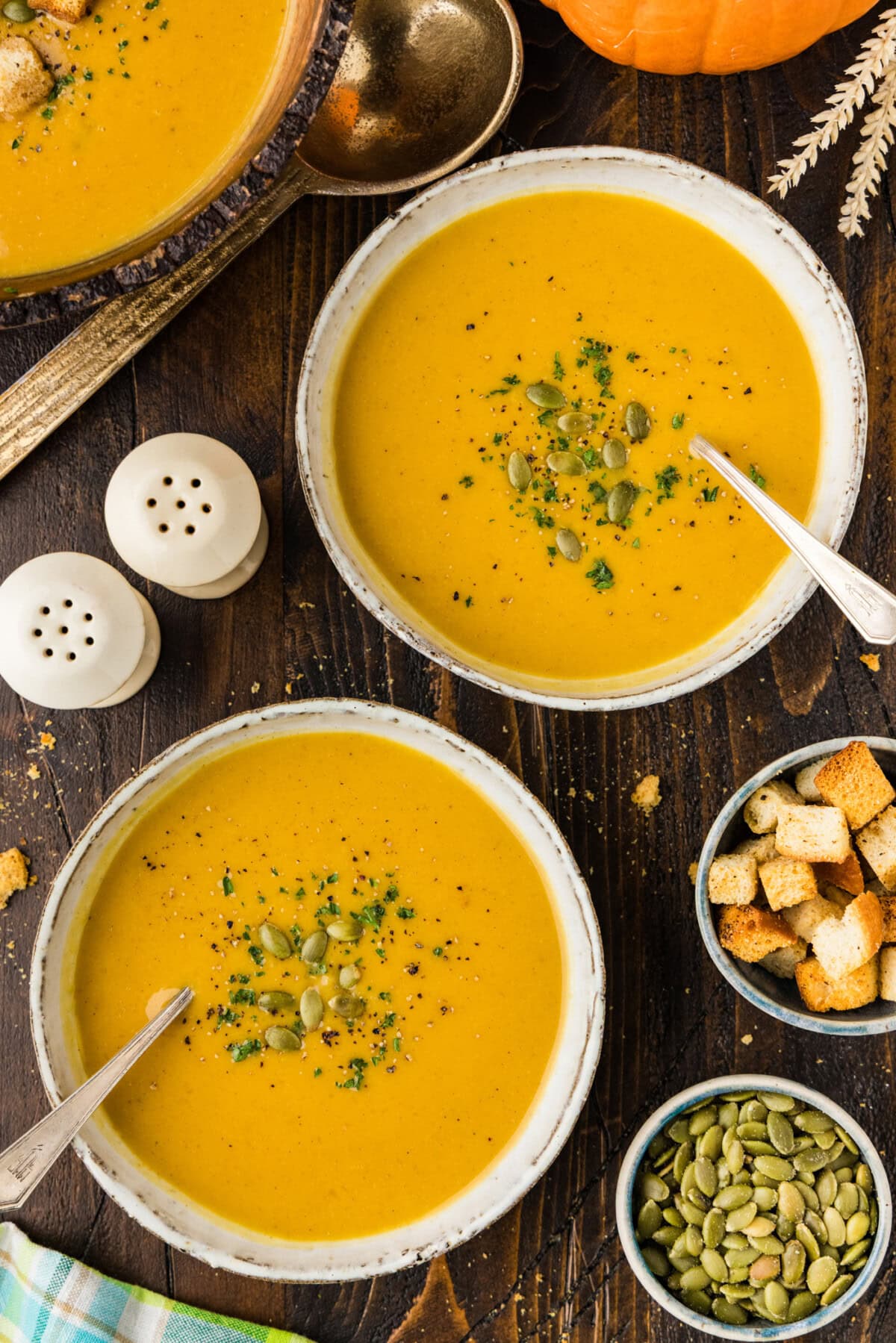 Two bowls of roasted pumpkin soup garnished with pepitas and parsley, served with small bowls of croutons and pumpkin seeds.