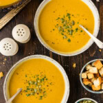 Two bowls of roasted pumpkin soup garnished with pepitas and parsley, served with small bowls of croutons and pumpkin seeds.