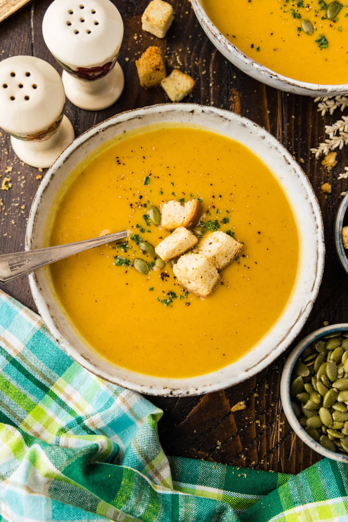 Bowl of roasted pumpkin soup topped with homemade croutons, pepitas and parsley, with a spoon and a plaid napkin.