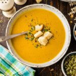 Bowl of roasted pumpkin soup topped with homemade croutons, pepitas and parsley, with a spoon and a plaid napkin.