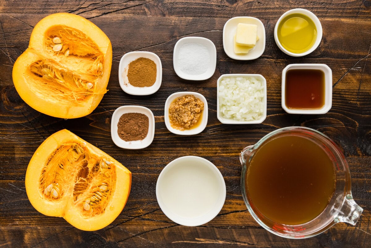 Ingredients for roasted pumpkin soup including a halved sugar pumpkin, olive oil, butter, roasted garlic, diced onion, cinnamon, nutmeg, maple syrup, heavy cream and vegetable broth.